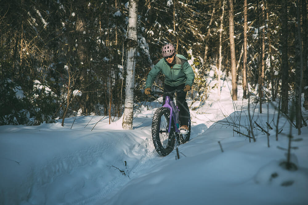A person riding a purple fat tire bike on a snowy forest trail, wearing a green jacket, dark pants, and a helmet.