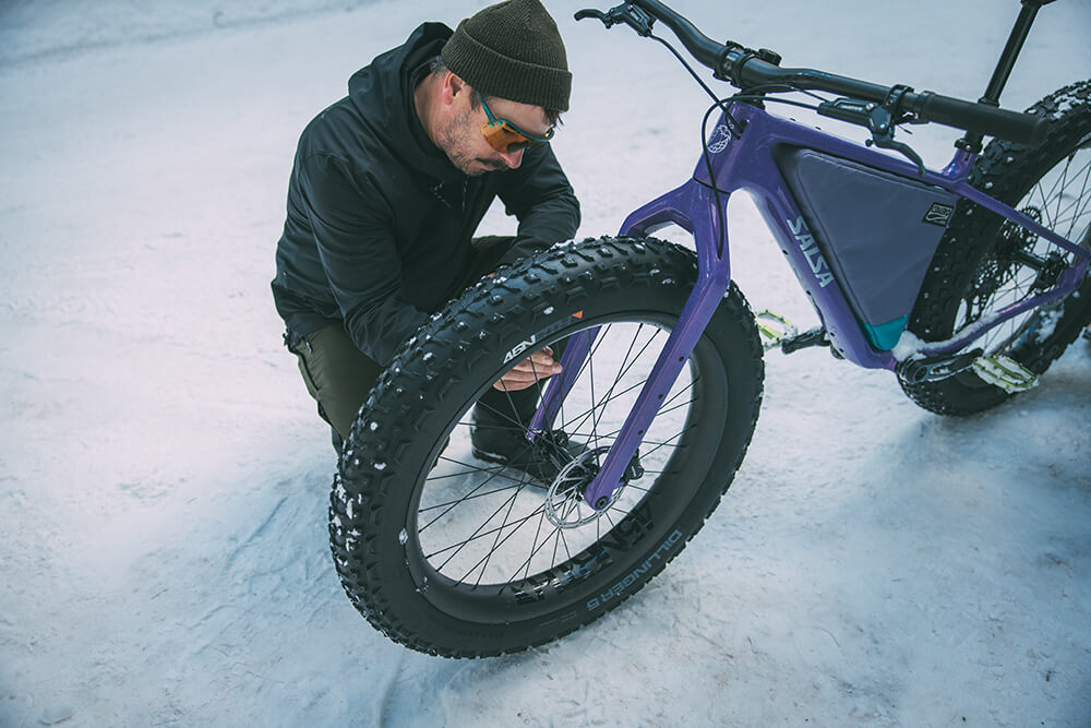 A man in winter clothing kneels beside a purple fat tire bike in the snow, inspecting the front tire.
