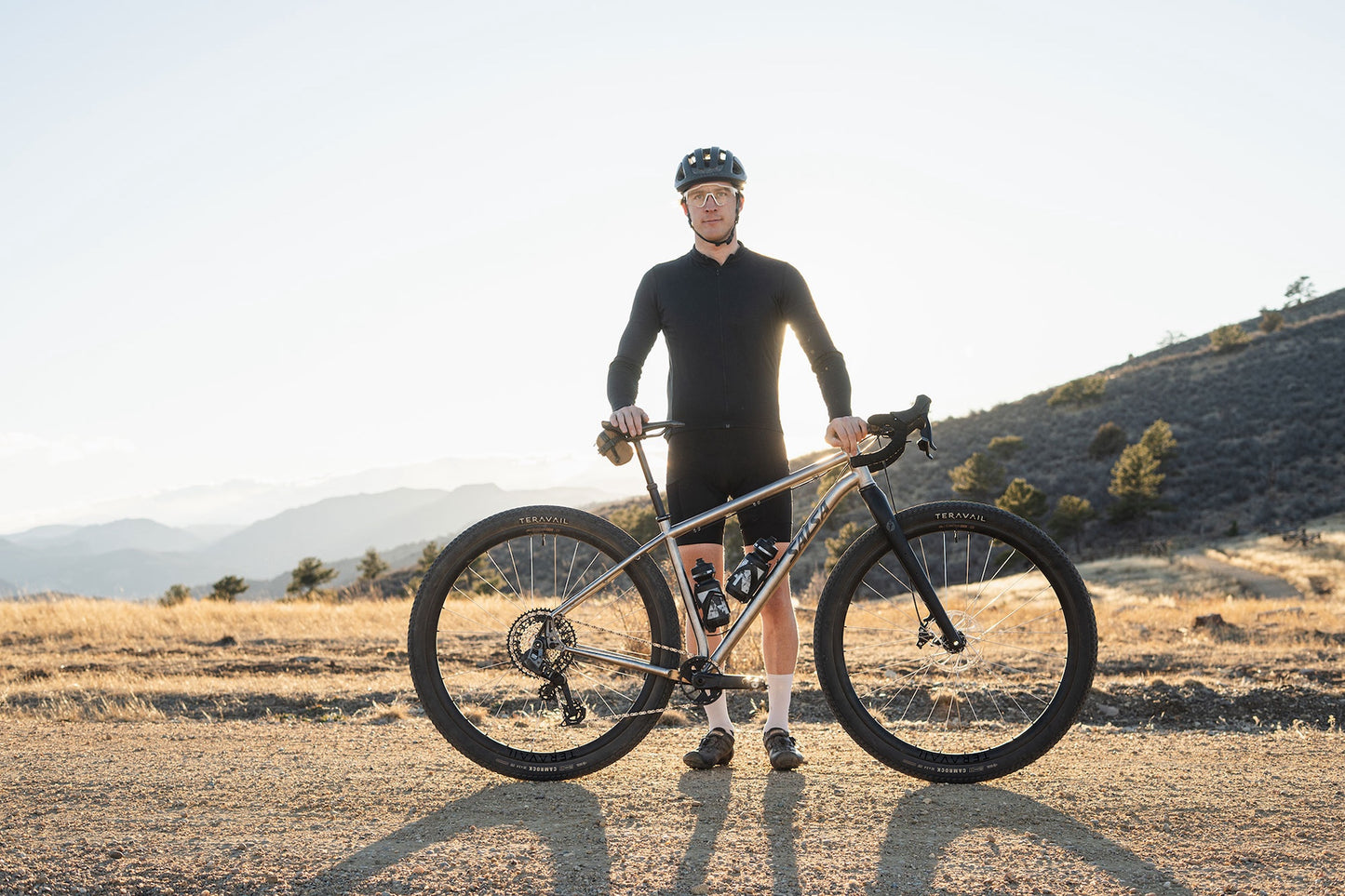 A gravel cyclist stands outdoors on a dirt road with a Fargo 32 Ti bike, wearing a black long-sleeve jersey, black shorts, and a black helmet, with dry grass and hills in the background.