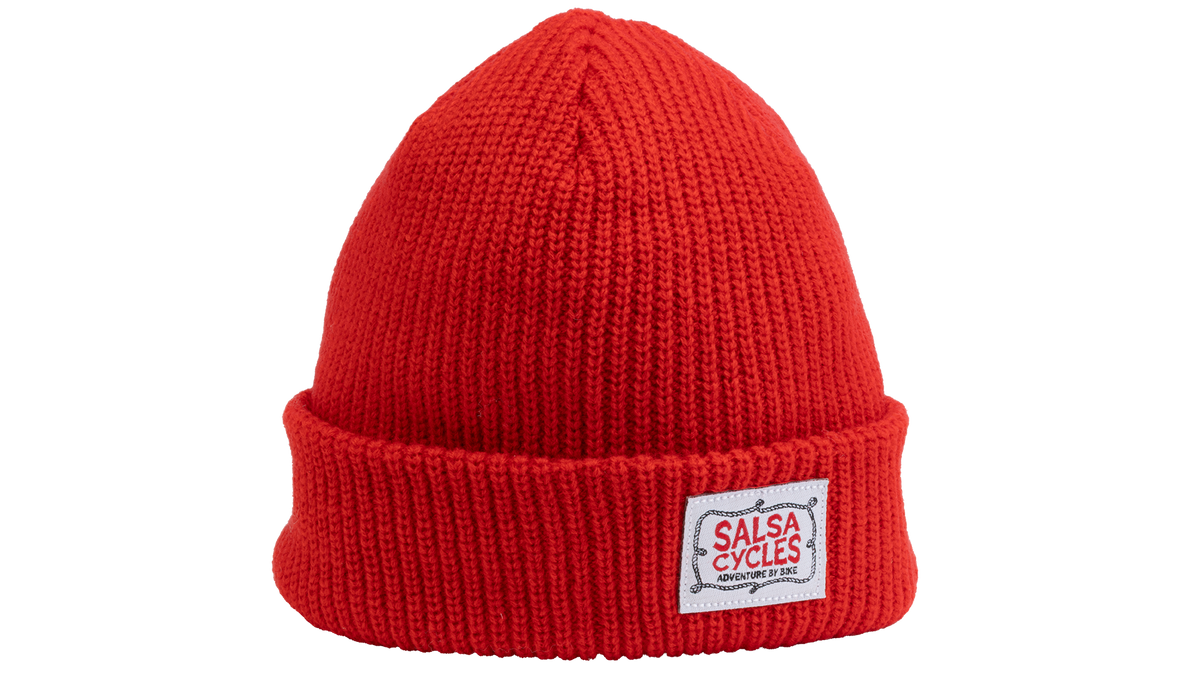 A bright red ribbed knit beanie hat with a folded cuff, featuring a white rectangular patch on the front that reads 'SALSA CYCLES'