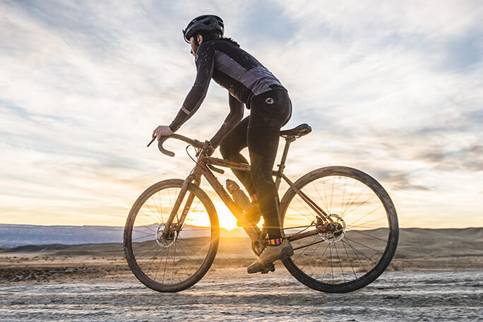 Cyclist on a drop bar bike rides on a barren gravel road with the sun setting in the background.