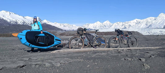 Pepper holding packraft next to campsite with two bikes parked, mountains in background