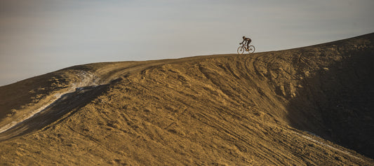 Left side view of a cyclist riding down a winding gravel and dirt trail along the spine of a bare hill on a clear day