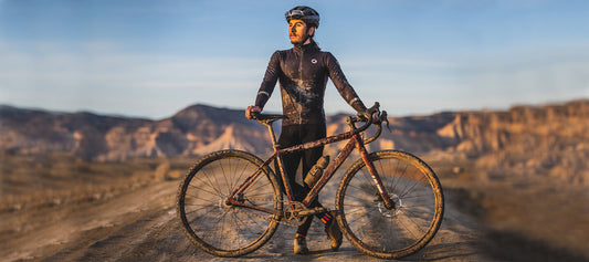 Front view of cyclist Leonardo Brasil standing on a gravel road with a muddy bike in front and rocky hills in background