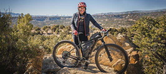 Smiling cyclist, Brooke Goudy, stands with a Salsa Cutthroat bike on a rocky mound in a brushy desert