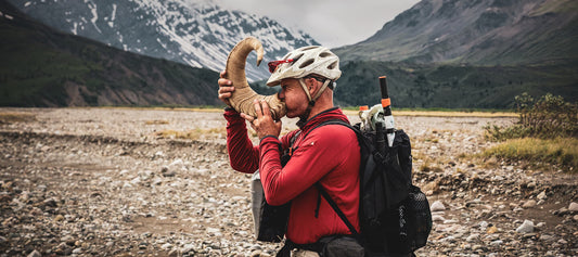 Left side view of a person with cycling helmet on, blowing air through a rams horn, in a mountain valley