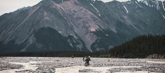 Front view of a lone cyclist, carrying their bike while walking through a shallow river, with mountains in the background