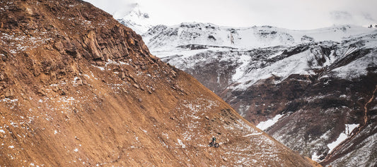 Downward, distant view of a cyclist walking their fat bike along a narrow, bare trail in the snowy mountains