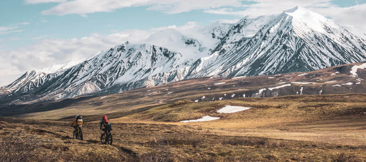 Rear view of 2 fat bike cyclists riding on a narrow dirt trail in a tundra field with snow covered mountains ahead of them