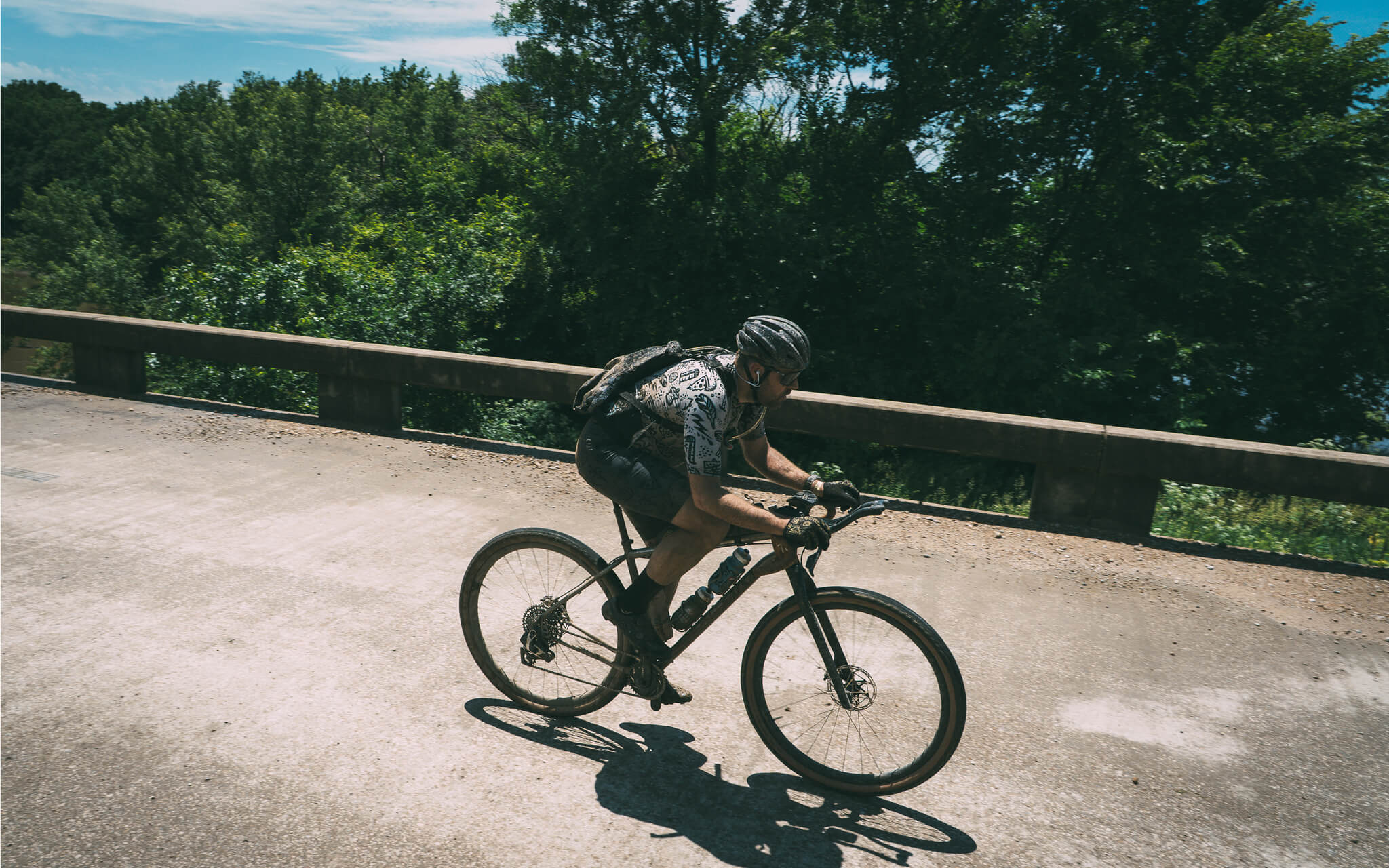 A cyclist wearing a helmet and backpack rides a Salsa Fargo 32 bike on a paved road with a cement guardrail and a dense green forest in the background.