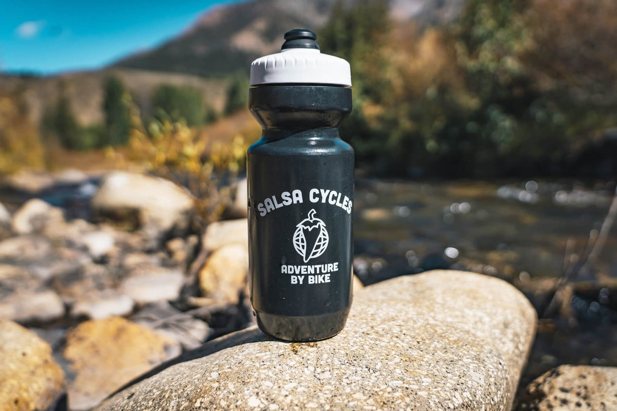 A black water bottle with a white lid stands on a large rock in an outdoor mountain stream setting. The bottle is printed with the white text 'SALSA CYCLES' and 'ADVENTURE BY BIKE' with a pepper globe logo.