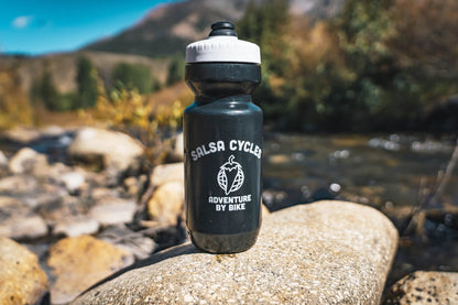A black water bottle with a white lid stands on a large rock in an outdoor mountain stream setting. The bottle is printed with the white text 'SALSA CYCLES' and 'ADVENTURE BY BIKE' with a pepper globe logo.