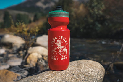 A red plastic water bottle with a green cap stands on a large rock outdoors near a stream, with blurred trees and hills in the background. The bottle has a white graphic of a cyclist and the text 'Salsa Cycles' and 'Send It Spicy!'.
