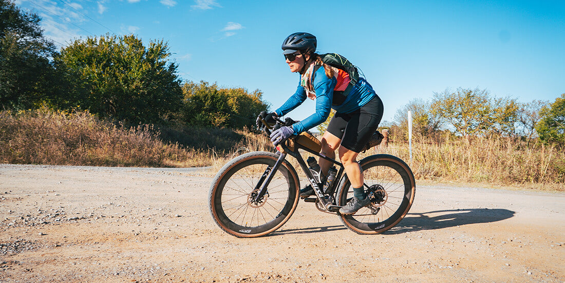 Cyclist riding a Salsa Flyway gravel bike on a dirt road with trees and clear sky in the background