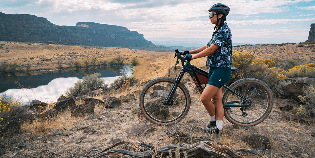 Person with a bicycle standing on a rocky trail with a scenic landscape in the background