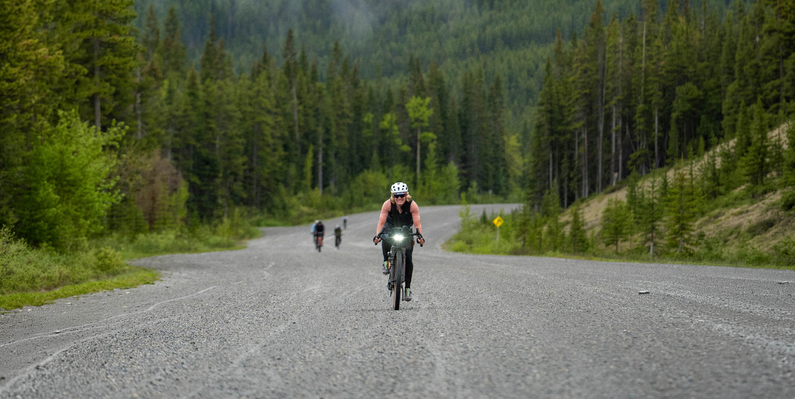 A cyclist rides along a gravel road surrounded by a dense green forest, with other distant riders on the road.