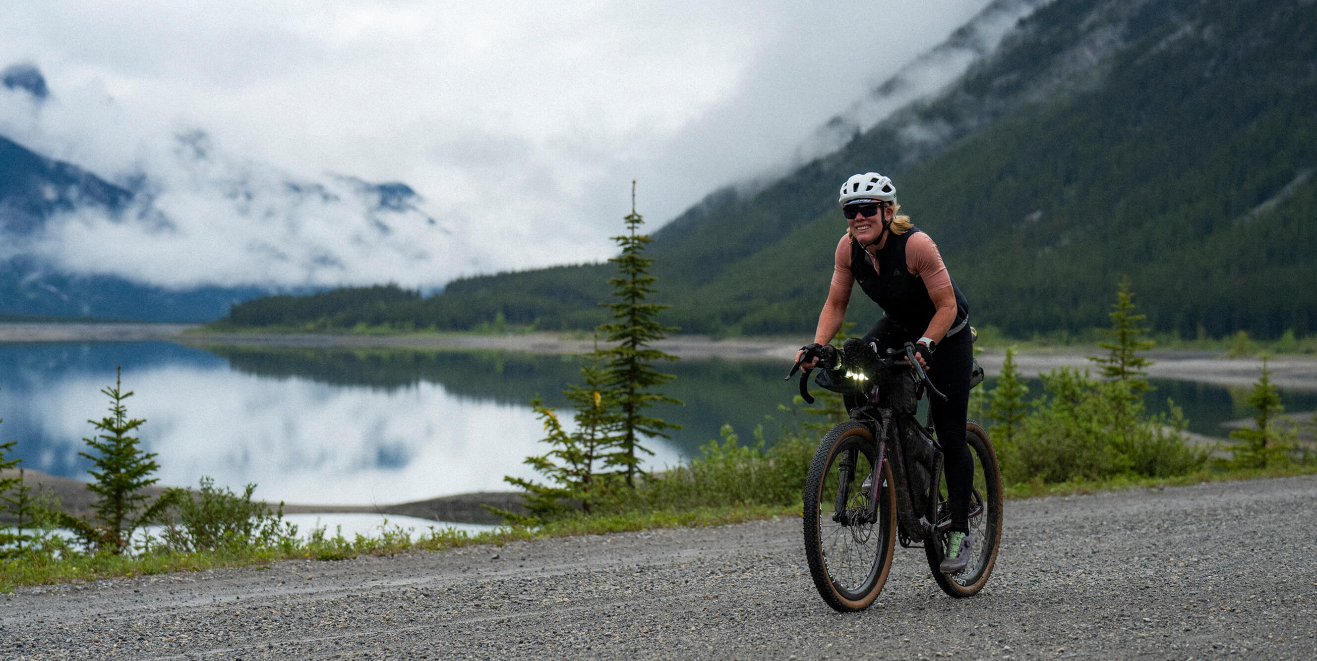 A cyclist wearing a white helmet and black outfit rides a Salsa Cutthroat bike on a gravel path beside a calm lake, with misty mountains and evergreen trees in the background.