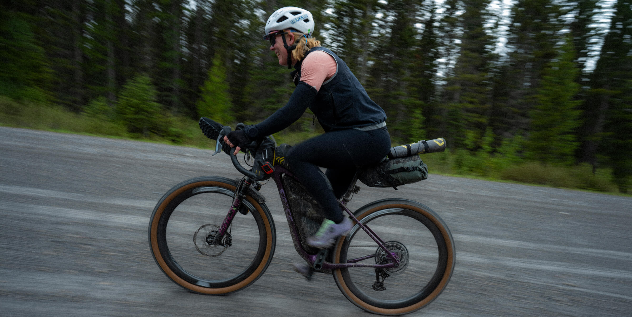 A person riding a Salsa Cutthroat bike on a paved road with a forest background. The rider is wearing a white helmet, black top, and black pants, and the bike has a rear rack with a bag.