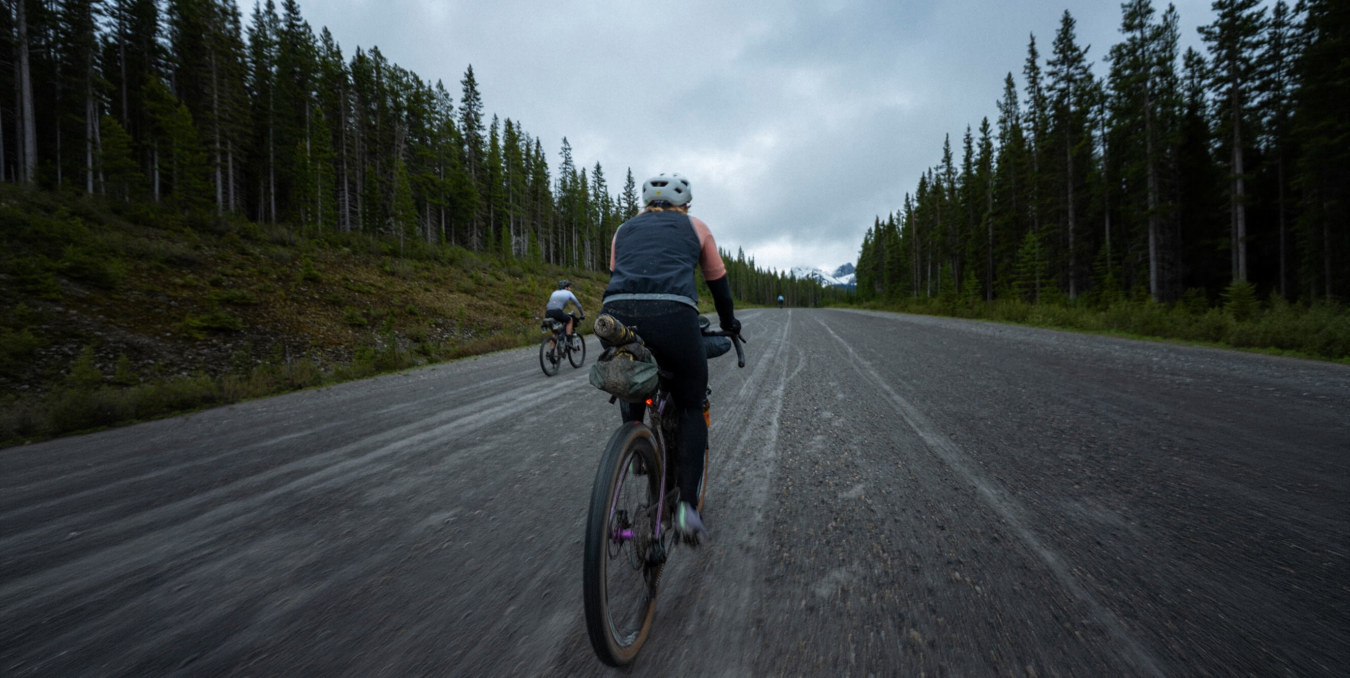 A cyclist rides away on a paved road between forested hills under an overcast sky, with another cyclist visible in the distance.