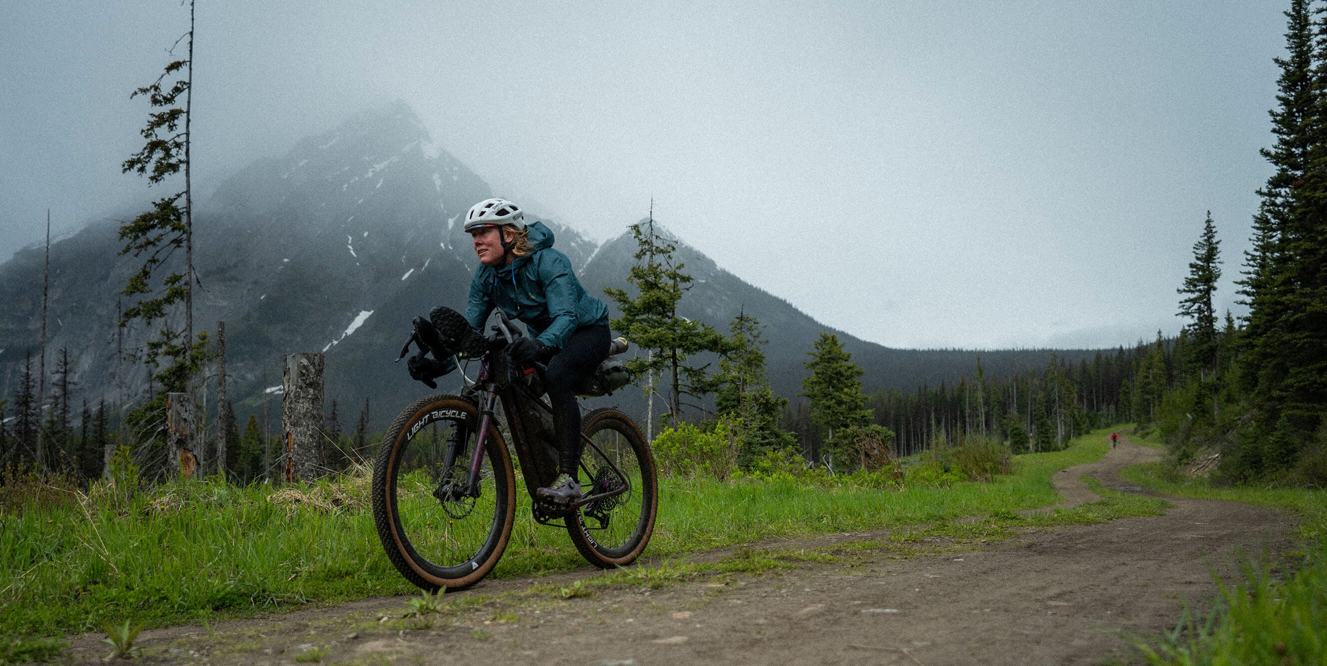 A person riding a Salsa Cutthroat gravel bike on a dirt trail in a misty mountain forest, wearing a teal jacket, black pants, and a white helmet.