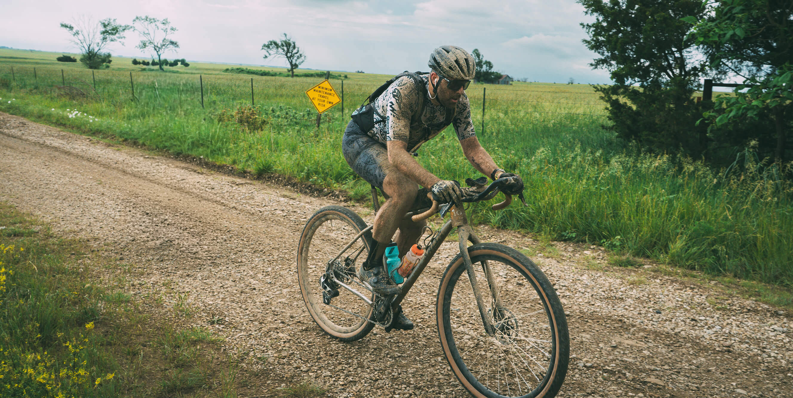 A cyclist riding a Salsa Fargo 32 gravel bicycle on a dirt path in a rural outdoor setting with grassy fields, trees, and a cloudy sky.