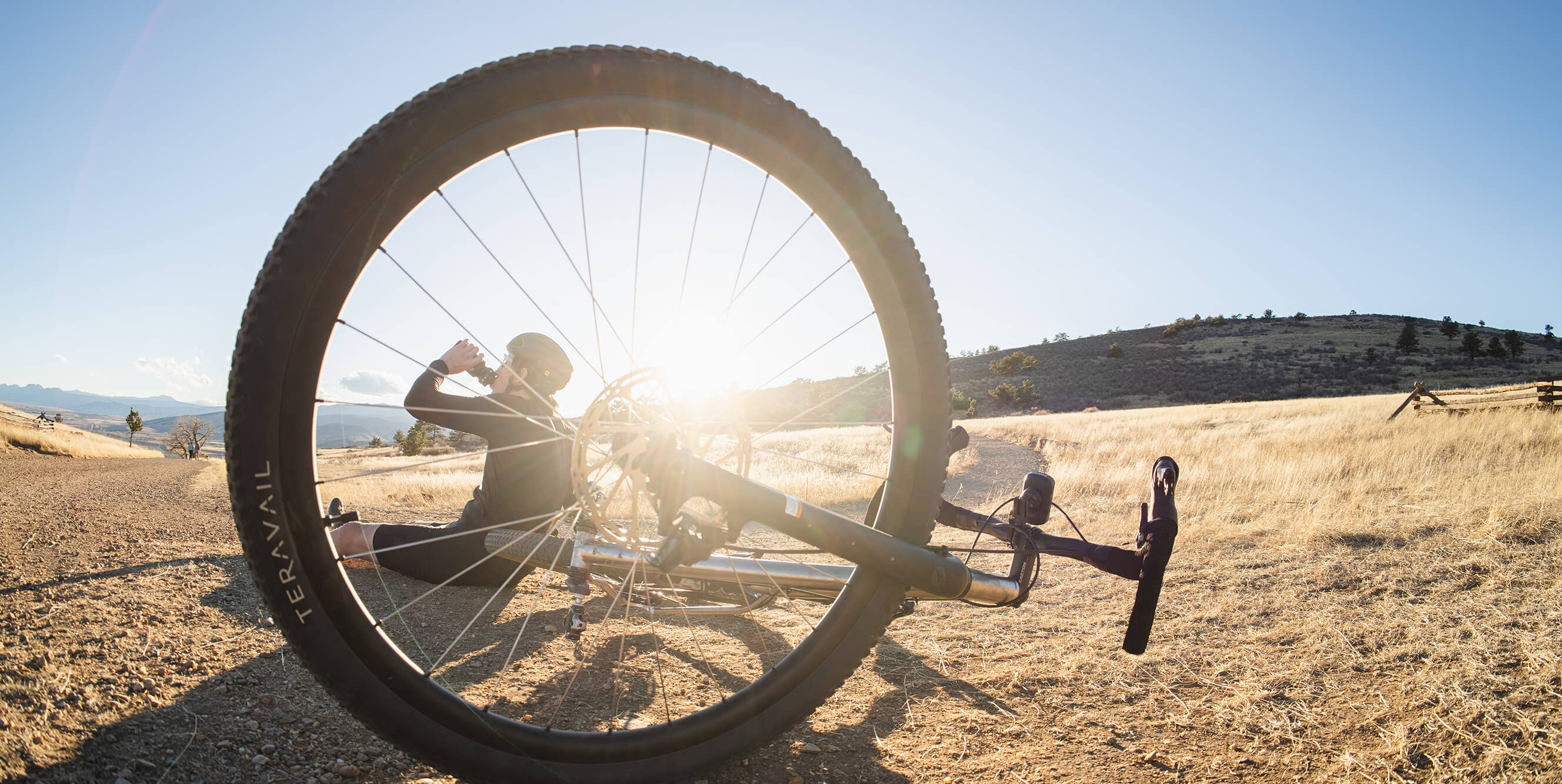 A Salsa Fargo 32 bike is lying on its side in an open grassy field, with a person sitting behind the large front wheel in bright sunlight. The background shows hills and a clear blue sky.