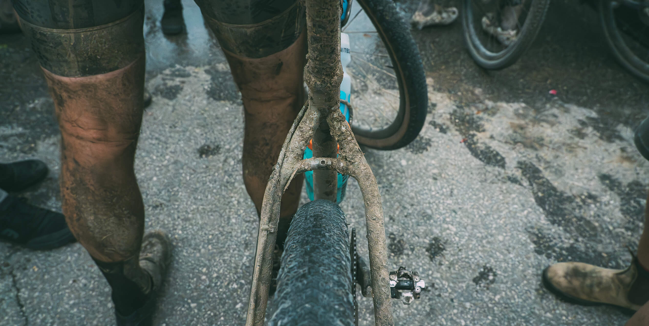 Close-up of a muddy Salsa Fargo 32 bicycle frame and rear wheel with a person's legs in muddy shorts standing beside it on a wet concrete surface.