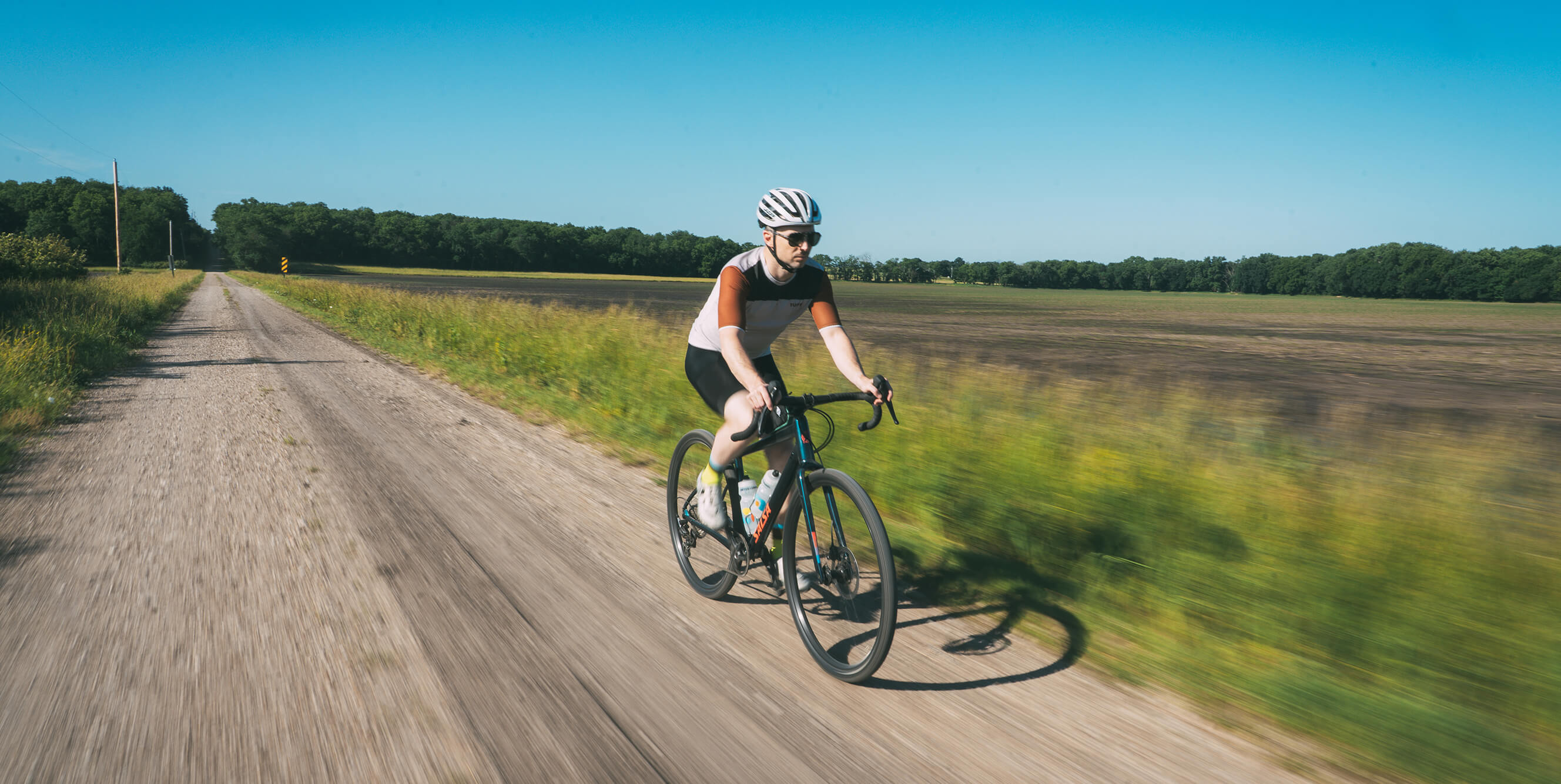 A cyclist rides a Salsa Journeyer gravel bike on a dirt rural road with green fields and trees under a clear blue sky.