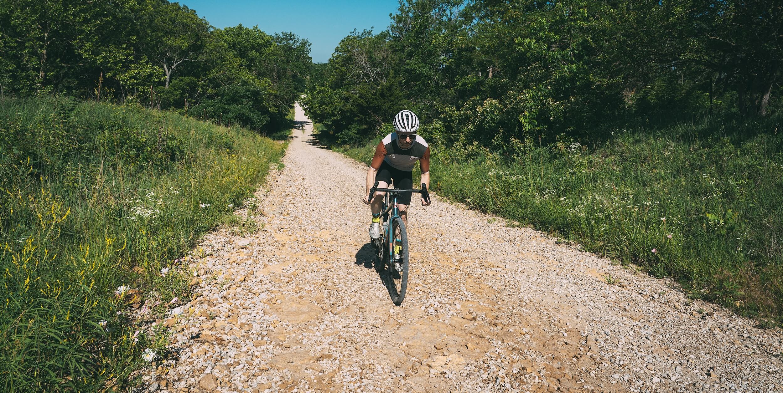 A cyclist rides away on a gravel path surrounded by green trees and grass under a blue sky.