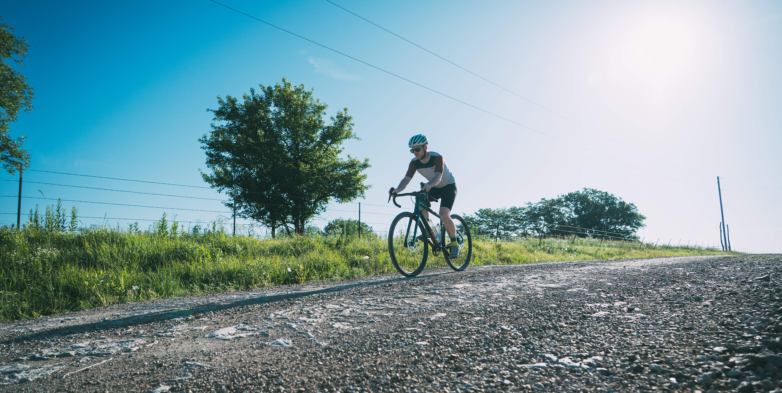 A cyclist rides a Salsa Journeyer gravel bike up a gravel path on a sunny day, with grassy fields, trees, and a bright blue sky in the background.
