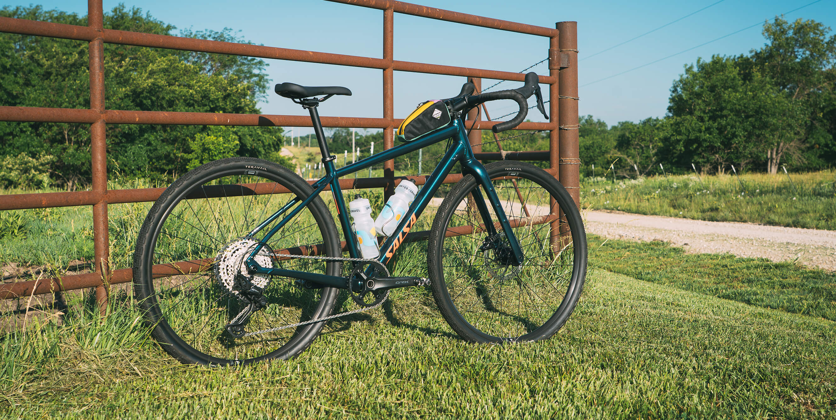 A dark teal Salsa Journeyer gravel bicycle with black components and a white water bottle mounted on the frame, parked outdoors on a grassy field next to a rusted metal fence.