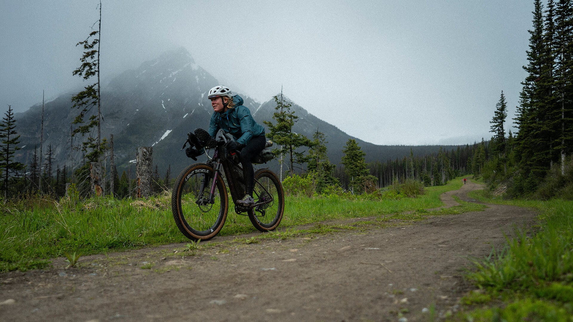 A person riding a Salsa Cutthroat on a dirt trail in a mountainous forest setting, wearing a teal jacket, black pants, and a white helmet.