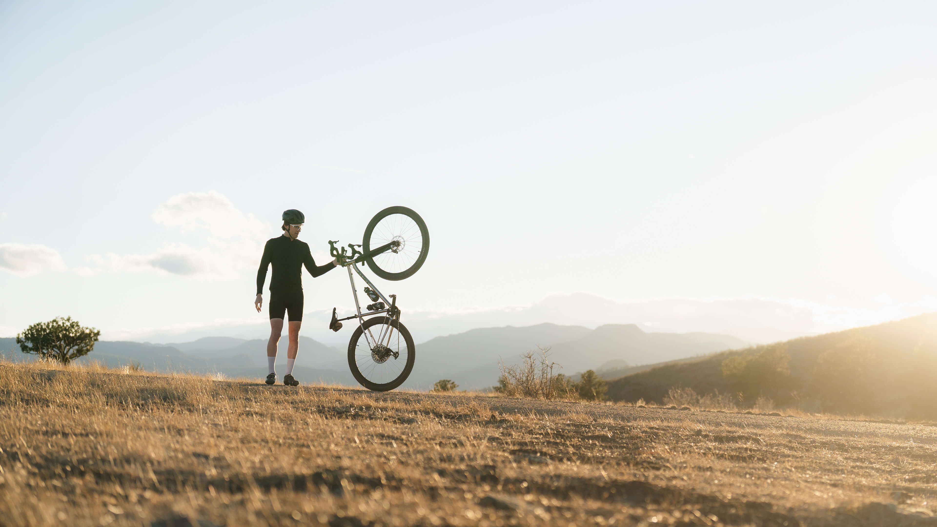Person carrying a bicycle through a scenic landscape with mountains and a clear sky.