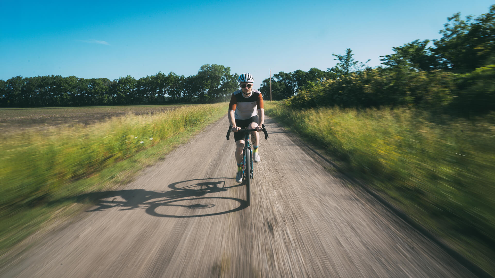 A cyclist rides down a dirt country road with grassy fields and trees on both sides under a clear blue sky.