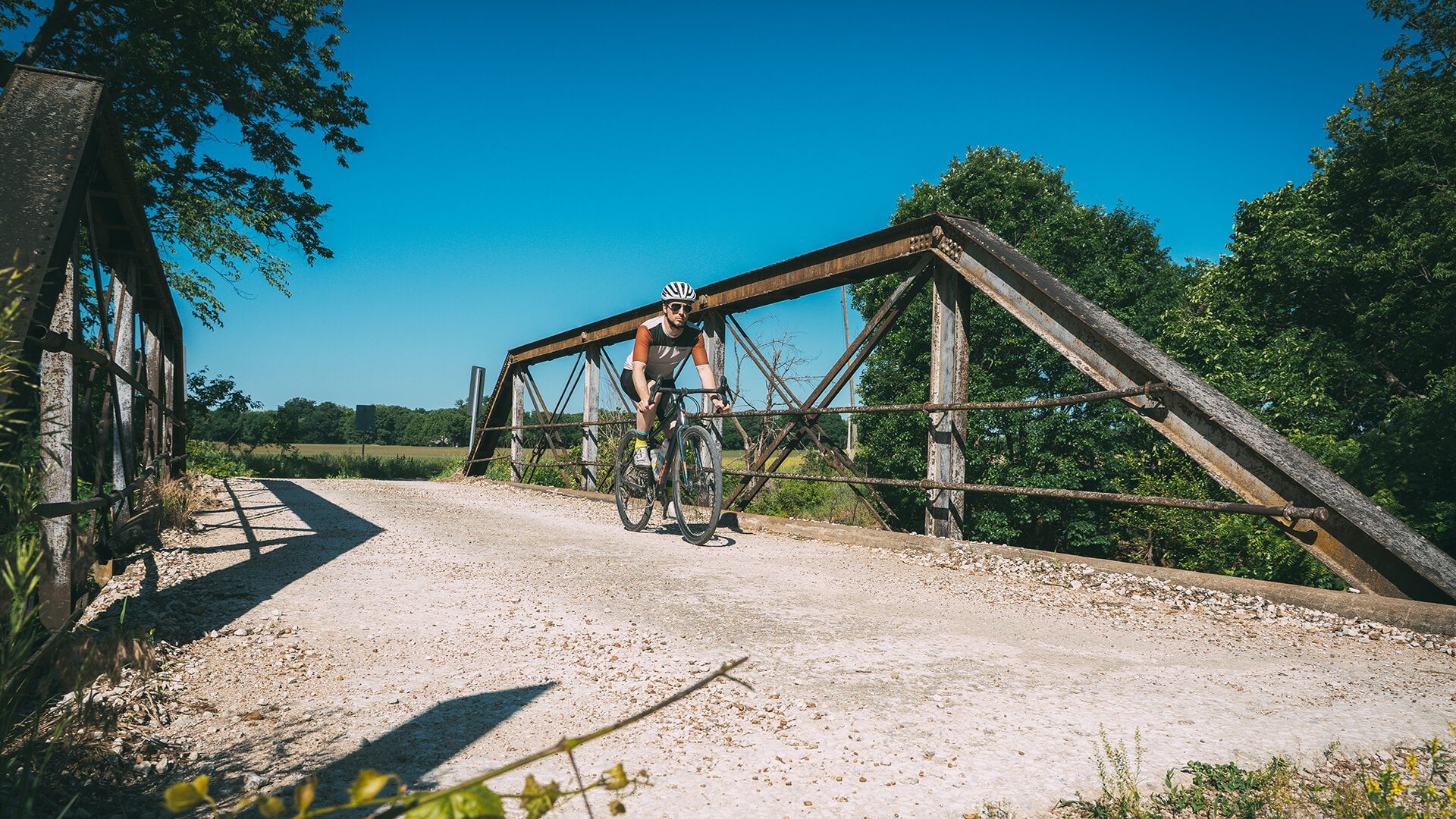 A cyclist rides a Salsa Journeyer gravel bike across a rustic wooden truss bridge on a dirt path, with trees and blue sky in the background.