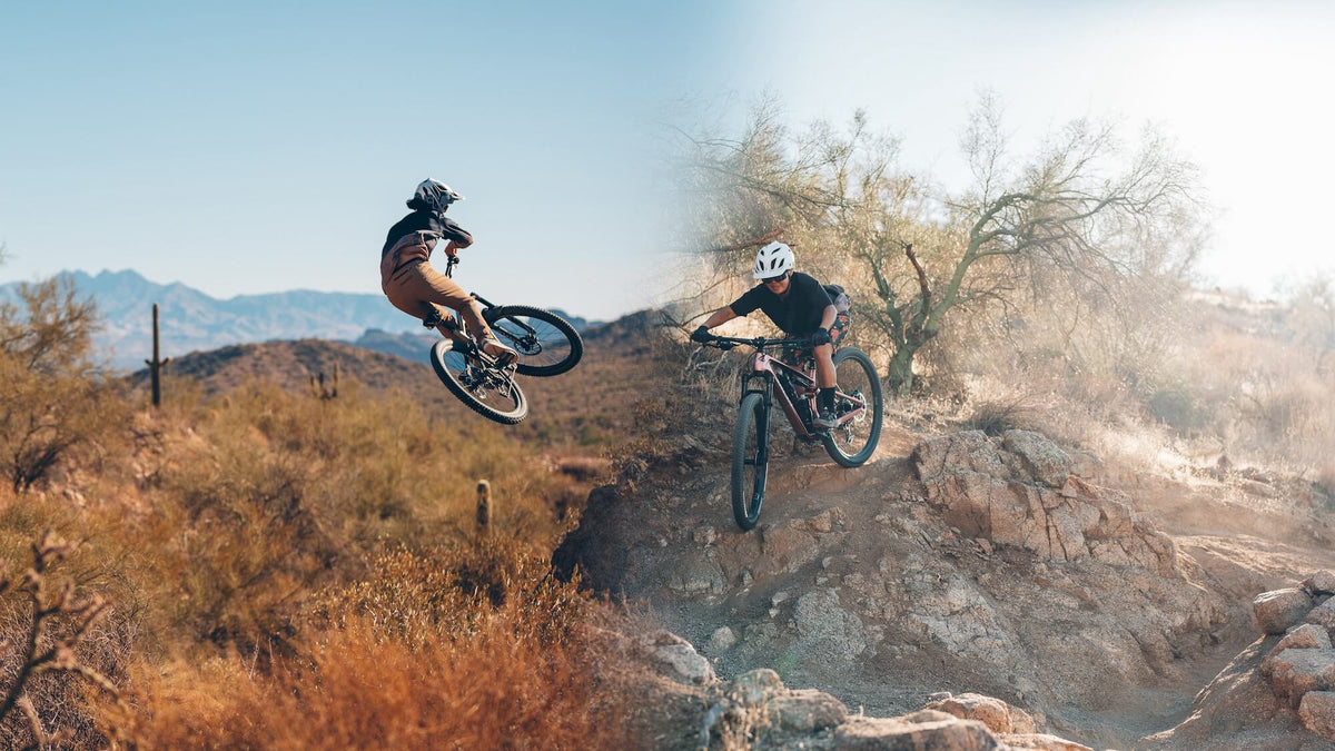 Two mountain bikers mid-jump on a rocky desert trail, with dry brush, trees, distant mountains, and a bright sky in the background.