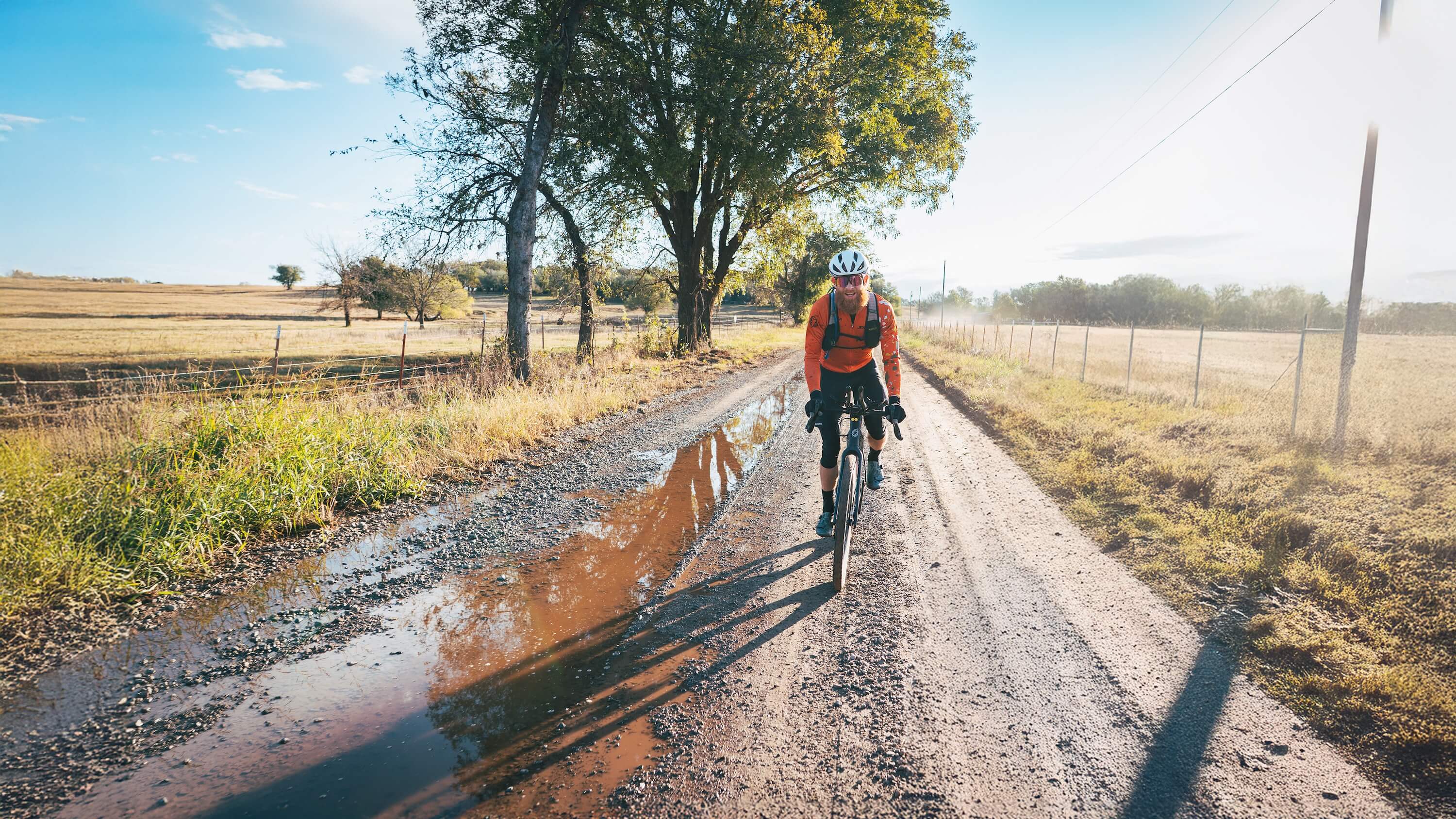 Cyclist riding a Salsa Flyway gravel bike on a rural dirt road with trees and fields in the background