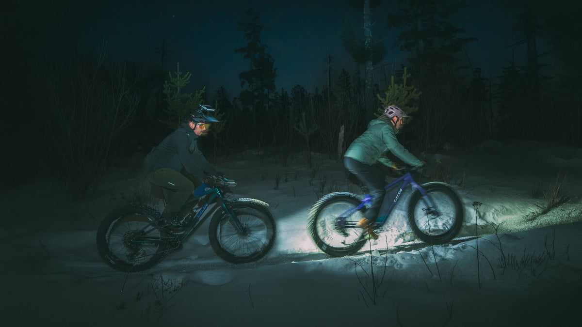 Two people riding fat-tire bicycles through deep snow at night in a dark forest, with headlights illuminating the path.
