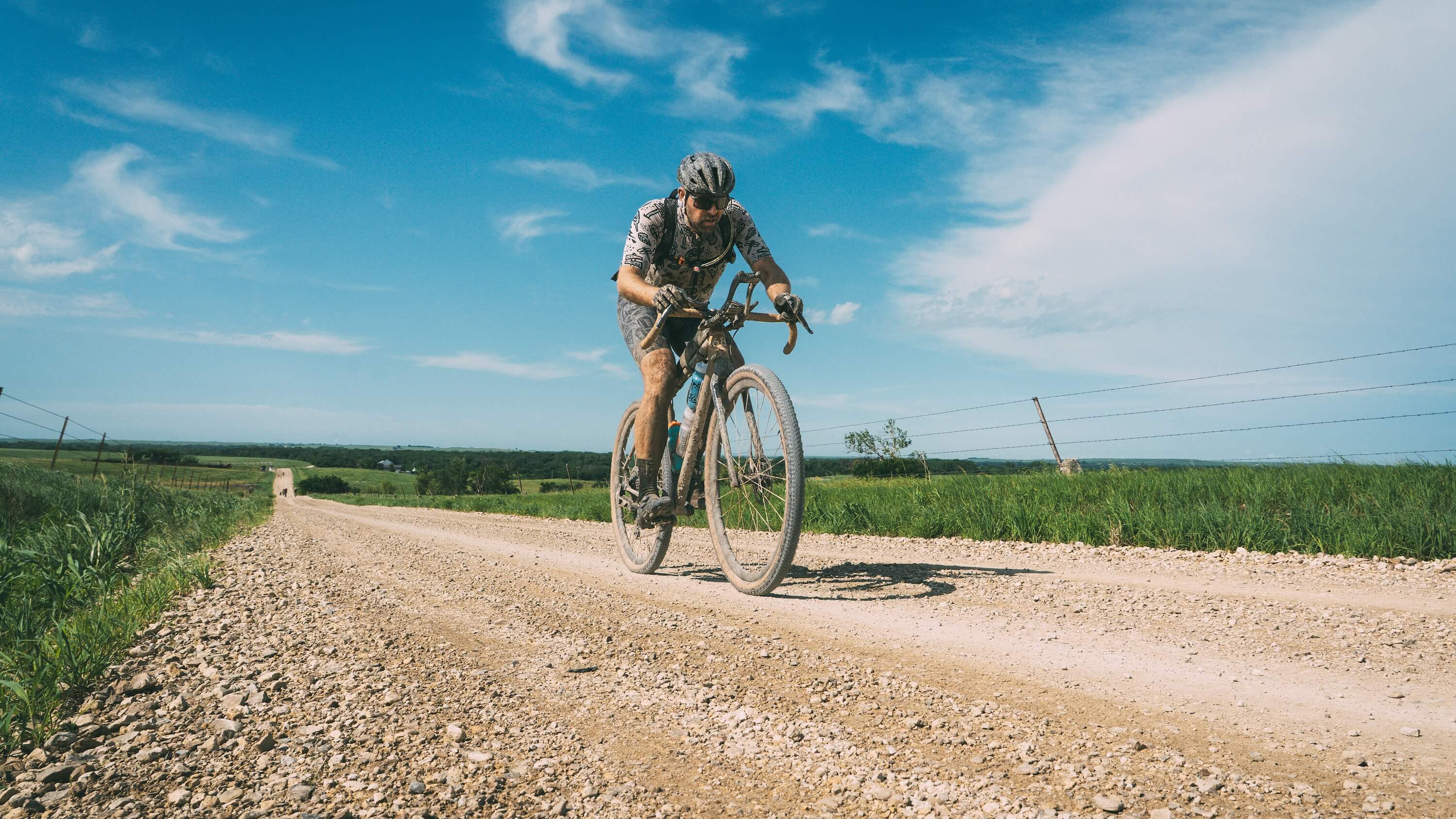 A cyclist rides a Salsa Fargo 32 gravel bike on a dirt road outdoors under a blue sky with clouds, with green fields on both sides.