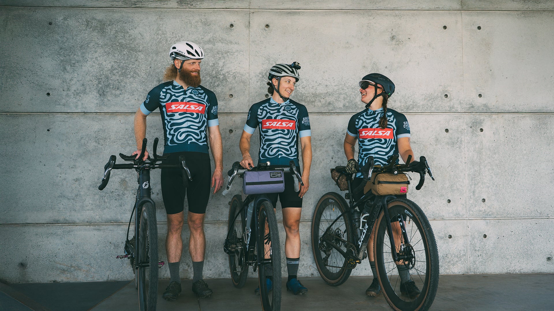 Three cyclists in matching Salsa cycling jerseys with bicycles against a concrete wall.