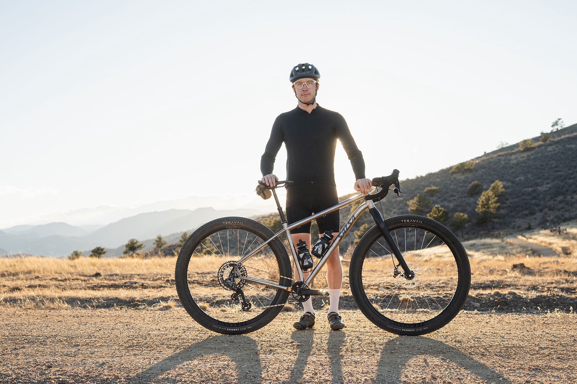 A gravel cyclist stands outdoors on a dirt road with a Fargo 32 Ti bike, wearing a black long-sleeve jersey, black shorts, and a black helmet, with dry grass and hills in the background.
