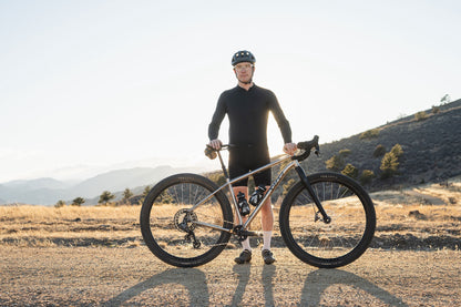A gravel cyclist stands outdoors on a dirt road with a Fargo 32 Ti bike, wearing a black long-sleeve jersey, black shorts, and a black helmet, with dry grass and hills in the background.