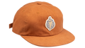 A rust-orange cotton flat-brim cap featuring a small embroidered patch of a pepper on the front