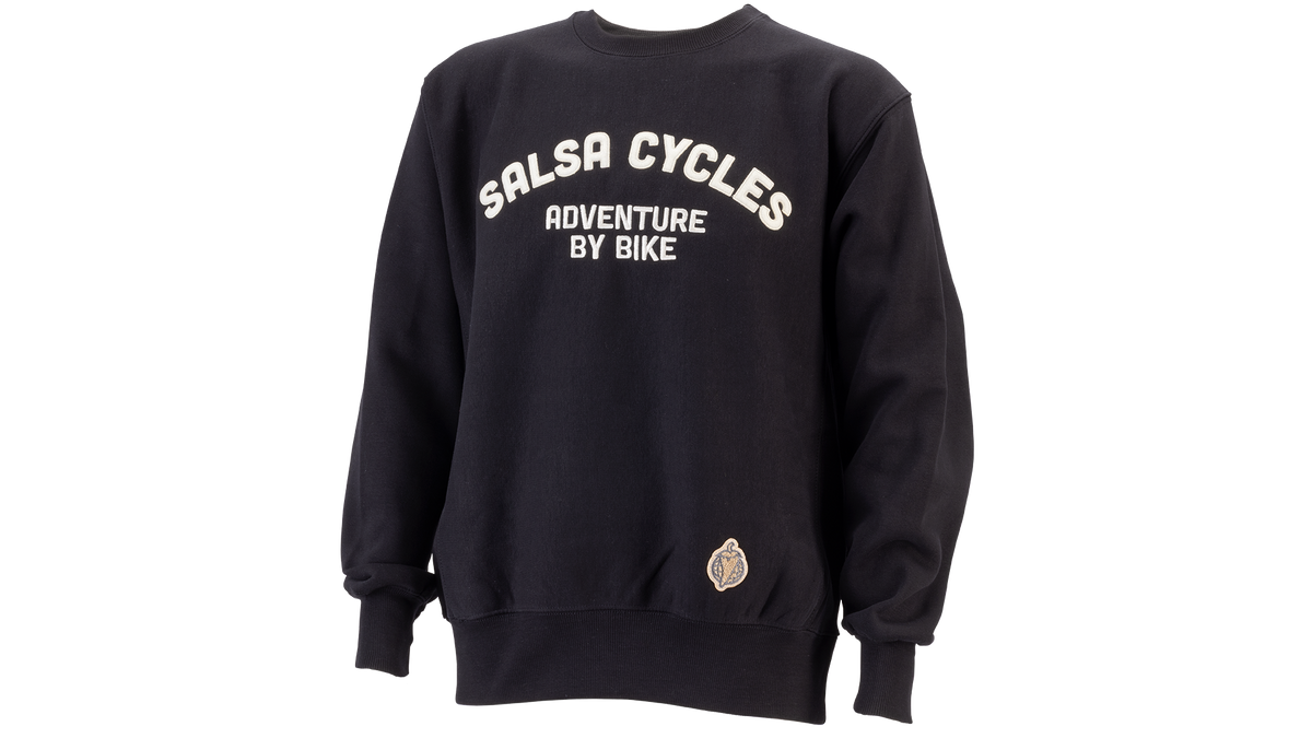 A black crewneck sweatshirt with 'SALSA CYCLES ADVENTURE BY BIKE' printed in white arched text on the chest, and a small circular patch near the lower left hem, shown on a plain white background.