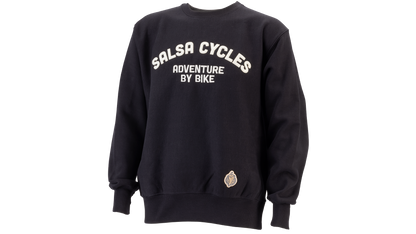 A black crewneck sweatshirt with 'SALSA CYCLES ADVENTURE BY BIKE' printed in white arched text on the chest, and a small circular patch near the lower left hem, shown on a plain white background.