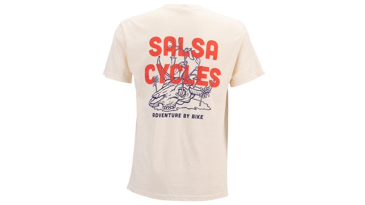 A cream-colored short-sleeve t-shirt shown from the back, featuring a large graphic print with the words 'SALSA CYCLES' in bold red letters and a blue line-art illustration of a person riding a bicycle. Smaller text below reads 'ADVENTURE BY BIKE'.