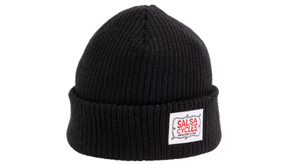 A black ribbed knit beanie hat with a folded cuff, featuring a white rectangular patch on the front with the red and white 'SALSA CYCLES' logo.