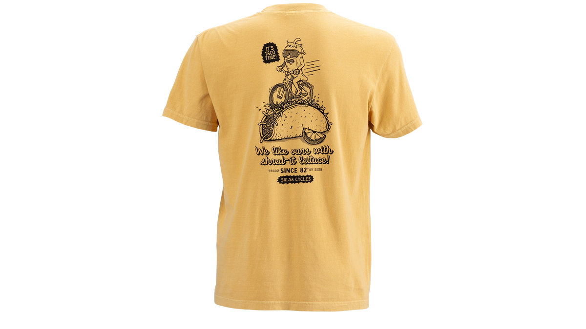 A yellow short-sleeve t-shirt shown from the back, featuring a large black graphic of a cartoon pepper riding a taco and the text 'We like ours with shred-it lettuce'.